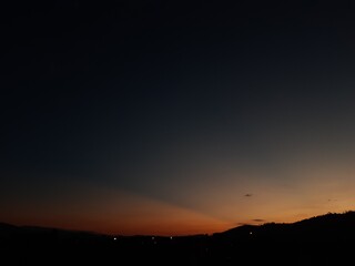 Un cielo despejado al atardecer, con un tono naranja del sol. Fondo un paisaje con montañas y luces de la ciudad.
