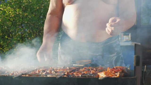 Fat funny man cooks kebabs and dances in the backyard. Unrecognizable man fries meat against the background of nature. The concept of gluttony and an idle lifestyle