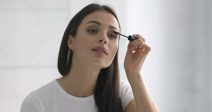 Confident happy young woman holding mascara apply on eyeleashes prepare getting ready in the morning, smiling beautiful girl doing make up put cosmetics on face looking in mirror in bathroom at home