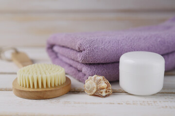 Obraz na płótnie Canvas A wooden brush for dry anti-cellulite massage with natural bristles, a seashell, a jar of moisturizer and a folded lilac towel on a white wooden background. Spa. Selective focus
