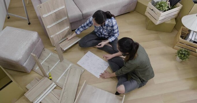 slow motion of two asian girls friends reading instructions to assemble wood bed in living room floor. women moving in new house apartment relocation lifestyle. pensive female roommates diy furniture