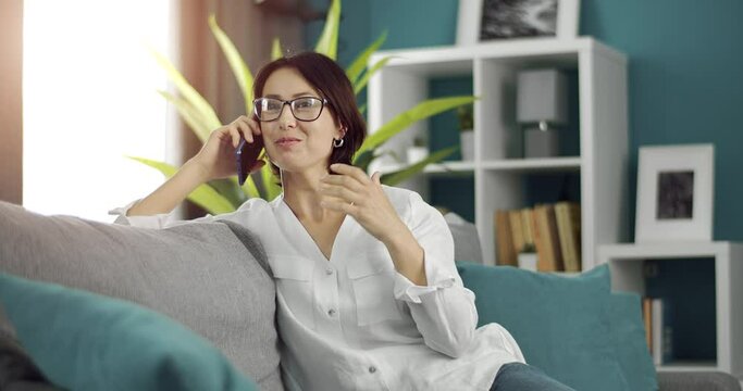 Happy woman in eyeglasses enjoying pleasant conversation on smartphone while sitting on grey couch. Maure lady with dark hair spending free time for chatting on modern gadget.