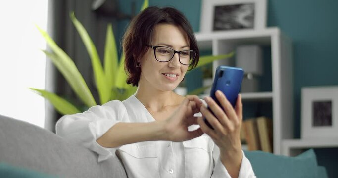 Smiling female in eyeglasses sitting on comfortable couch and using modern application on smartphone for convinience. Concept of people, home and technology.