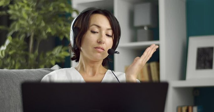 Charming woman in white shirt using laptop and headset for video conversation while sitting on couch. Concept of modern technology amd remote work.