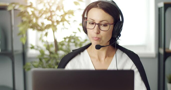 Smart mature worker wearing headset during video conference on laptop while sitting at table. Woman in formal clothing working at modern office center.