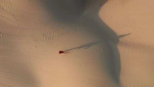 A young female in a beautiful fluttering red dress walking at the natural desert landscape. 4K Aerial drone view of a woman making footprints on the sand dune at sunset time. People video, California