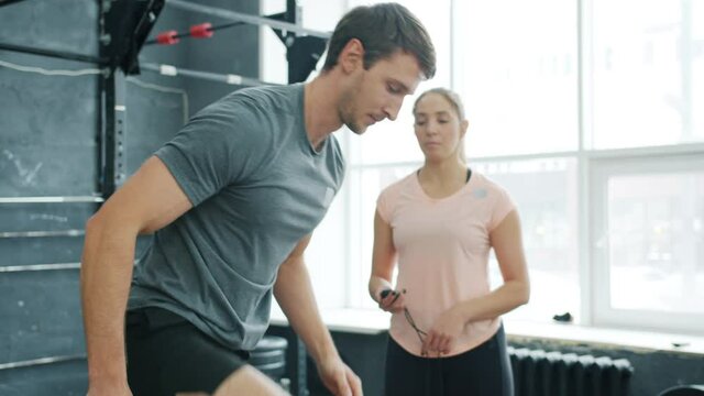 Handsome guy in sportswear and sneakers is jumping on wooden box training with crossfit instructor in gym focused on exercise. People and sports concept.