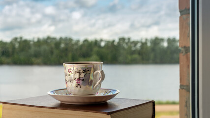 Cup of coffee and saucer on an old book on a background of window with blurred landscape