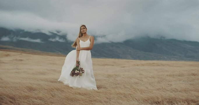 Young beautiful woman in windy field by the mountains with flowers in slow motion, bohemian white dress bouquet