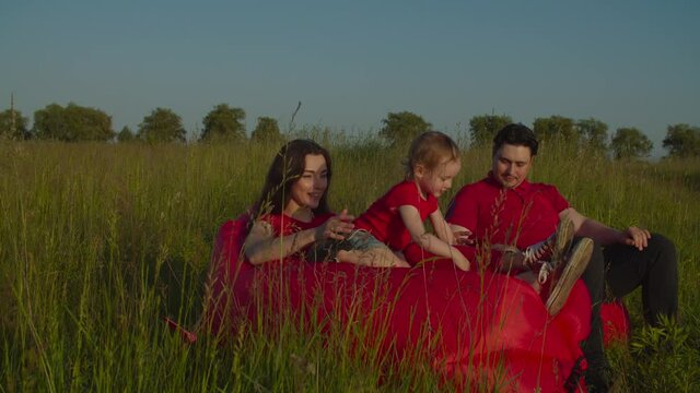 Carefree happy family with cute playful baby girl sitting on beanbag chairs on green field, lounging, having fun and bonding while spending leisure in summer nature during vacations in countryside.