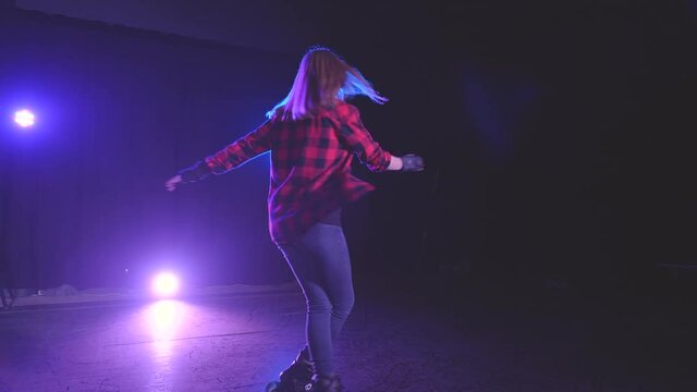 Young attractive girl rollerblades on stage illuminated by spotlights of abandoned building and spins on front wheels with crossed legs. Female roller skater shows cool performance in dark studio.