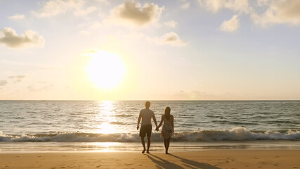 happy couple walking joining hands along beach to ocean surf against sunset at tropical resort close up