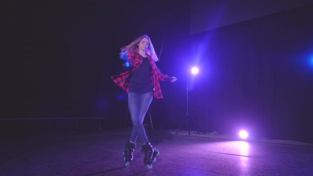 Blonde roller girl with protective gloves, gracefully swirls on rollerblades on front wheels in dark studio under neon pink blue light of spotlights in slow motion. concept active sports lifestyle