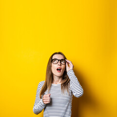 Young woman looks up in surprise, holding on to glasses on a yellow background.