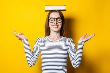 Foto op Canvas Young woman meditates with closed eyes holds a book on her head on a yellow background. © Alex