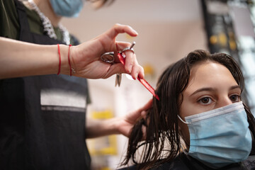 Hairdresser and girl child customer in a salon with medical masks during virus pandemic. Working with safety mask.