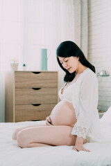 sexy fashion asian chinese woman looks at naked pregnant belly sitting in white comfort bed. young motherhood mom relaxing in bedroom while one hand holding bare tummy careful take care unborn baby