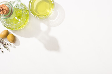 top view of olive oil in bowl and bottle near green olives, herb on white background