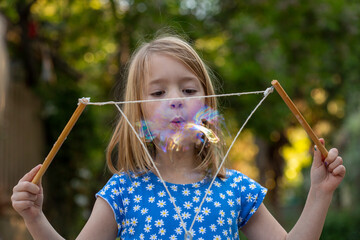 Young girl in a blue and white dress, blowing large bubbles in a backyard setting
