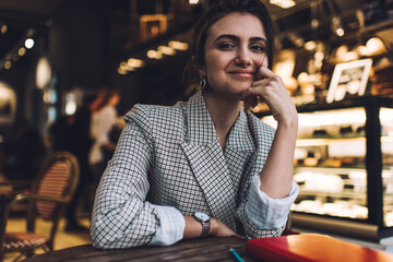Portrait of pretty Caucasian student holding literature best seller in hand and smiling at camera, charming Caucasian reader enjoying pastime with hobby posing during leisure in cafe interior