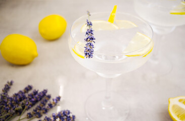 Sparkling water with ice and lemon in a margarita glass stands on a gray concrete background. The glass is decorated with a slice of lemon and lavender. Near are two lemons and lavender flowers.