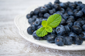 Juicy and fresh blueberries on a white plate with green mint leaves on a light wooden background. Blueberry antioxidant organic superfood in a bowl concept for healthy eating and nutrition. Close-up