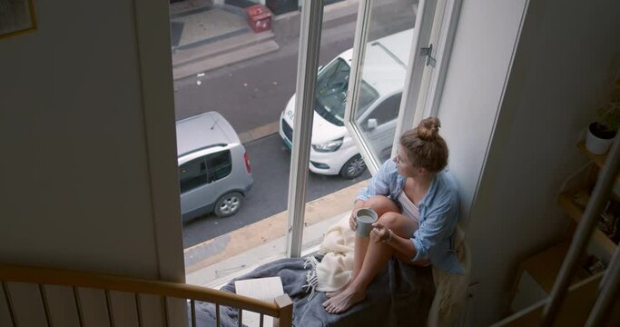 Cosy and comfortable, wrapped in blanket young woman enjoys cup of hot tea or coffee on balcony. Lazy sunday morning, weekend relaxation. Self contemplation concept. Look out of window on street