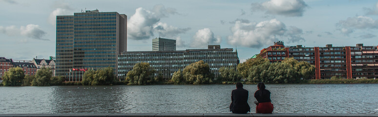 Panoramic crop of people sitting on promenade near canal with buildings and cloudy sky at background, Copenhagen, Denmark