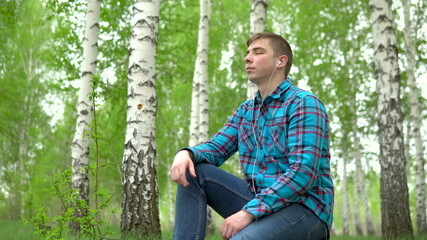 A young man sits in nature with headphones in his ears. A man sits on a stump in a birch forest and...