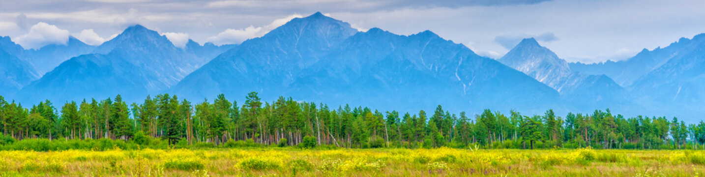 Panoramic summer landscape of mountain range with peaks, valley with green grass, grove and cloudy sky. Natural background with space for text. Eastern Sayan, Tunka National Park, Buryatia, Russia