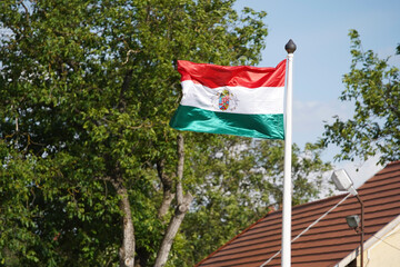 Flag of Hungary on the mast in the wind. Blue sky, a tree and a red roof background. Europe Hungary