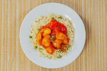 Serving of Chinese style sweet and sour chicken with rice on a white plate and bamboo table. Top view.