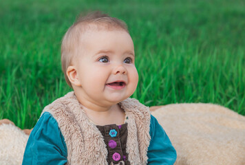 toothless baby in a fluffy multi-colored dress sitting on a bedspread on green grass and laughing