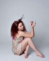 full length pose of red hair girl wearing floral fairy dress and headdress. sitting pose on grey...
