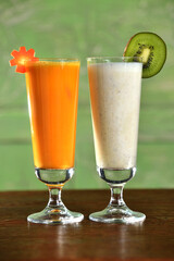 Two smoothie cocktails (kiwi fruit and vegetable carrot) in tall glasses on green background set on wooden table