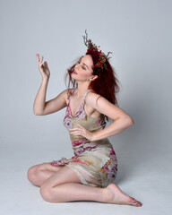 full length pose of red hair girl wearing floral fairy dress and headdress. sitting pose on grey studio background.