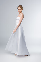 Fototapeta na wymiar smiling bride in elegant wedding dress standing on grey