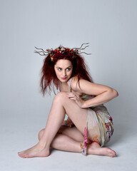 full length pose of red hair girl wearing floral fairy dress and headdress. sitting pose on grey...