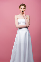 Fototapeta premium surprised bride in white wedding dress touching face isolated on pink