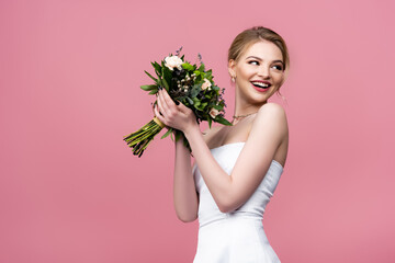 cheerful girl in white wedding dress holding flowers and looking away isolated on pink