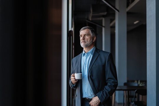 Mature businessman drinking a coffee and looking out of a window at the city from an office building.