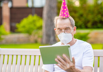 Old man wearing party's cap and protective mask celebrates his  birthday with his family on video call during the coronavirus epidemic