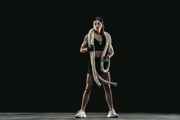 Beautiful young female athlete practicing on black studio background, full length portrait. Sportive fit brunette model with ropes. Body building, healthy lifestyle, beauty and action concept.