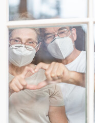 Elderly couple wearing protective face masks watch through their home window and show heart sign together during the coronavirus epidemic