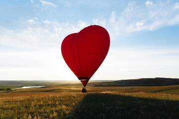 Fototapeta premium hot Red air balloon heart shape flying into sunset over valley landing or taking off