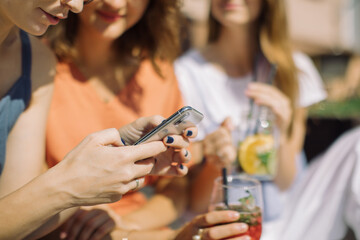 Girls sitting on summer terrace in the city, looking at mobile phone screen, drinking lemonade, talking. City life concept.