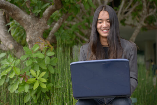 Happy young Asian businessman with long hair using laptop at the park