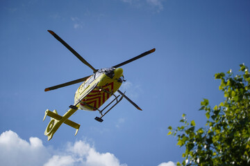 Fototapeta na wymiar The air ambulance, service arrives at the scene of an accident. Flying yellow medical ambulance helicopter over the trees. Background of blue sky with clouds. Pest county / Hungary - 06/01/2020