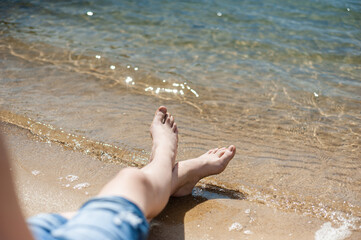 The teenager's legs on the shore near the surf line. Turquoise water and yellow sand. Summer holidays by the sea.