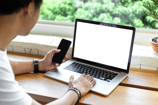 A close-up view of the back of a man using communication technology devices such as laptops and mobile phones on a wooden table abasing glass window and  tree. communication technology concept.